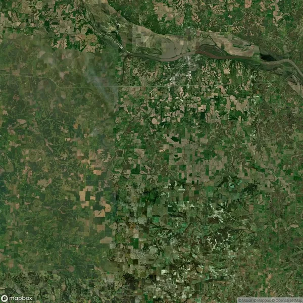 Aerial view of a patchwork landscape with fields, roads, and vegetation in varying shades of green and brown. A river flows diagonally through the top section of the image.
