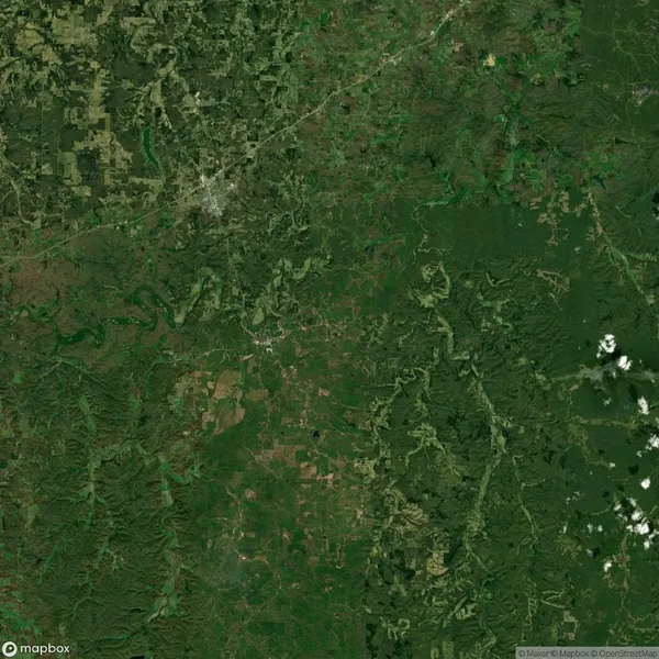 Aerial view of a densely forested rural landscape with roads, rivers, and patches of cleared land. A small urban area is visible in the upper left section. Sparse clouds in the lower right.