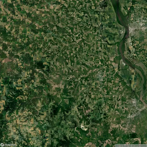 Satellite view of a rural landscape with patches of forest and agricultural fields. A river flows along the right side, with a cluster of urban development visible in the upper right corner.