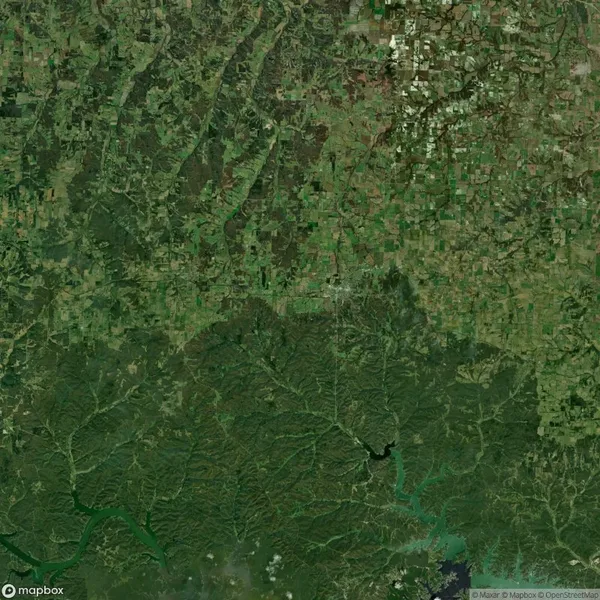 Aerial view of a landscape with irregular green patterns, showing agricultural fields and patches of forested areas. Roads and small settlements are scattered throughout.