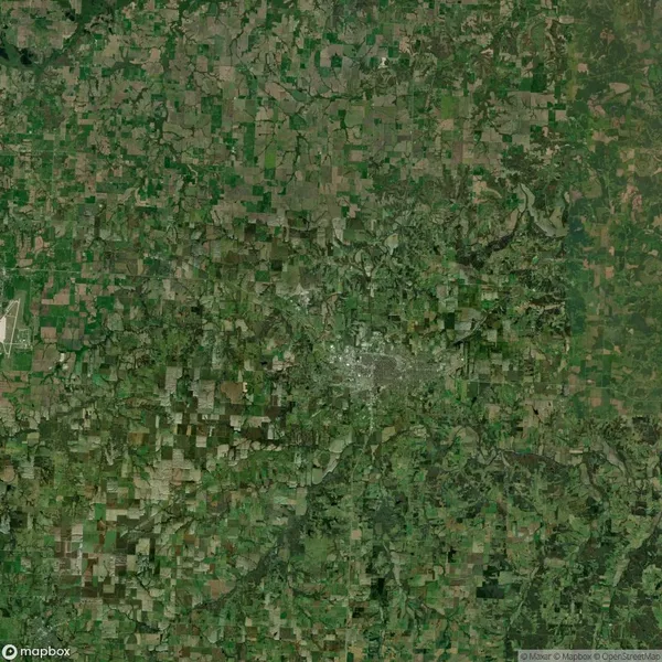 Satellite view of a rural landscape with a patchwork of green and brown fields, intersected by roads and waterways. A denser, possibly urban area is visible in the center.