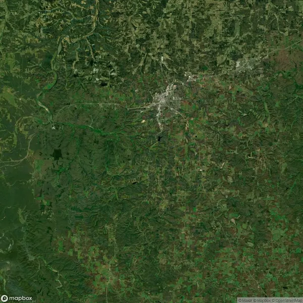 Satellite view of a region with dense green forested areas, rough terrain, and a clustered urban area evident in the top center.