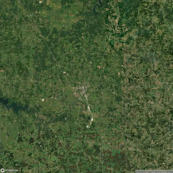Aerial view of a rural area with scattered green fields, patches of forest, and a small town center surrounded by agricultural land. Roads and a river are visible.
