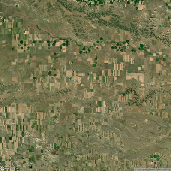 Aerial view of patchwork farmland featuring rectangular fields and circular crop patterns in shades of green and brown. Sparse greenery and dirt roads visible.