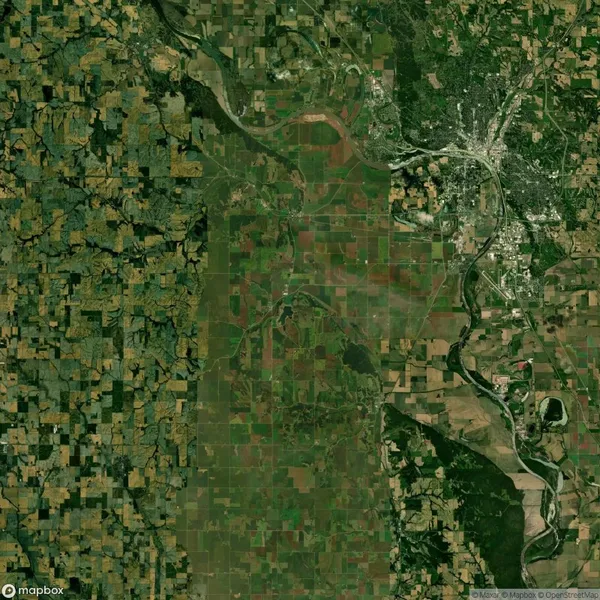 Satellite view of a landscape showing patchwork fields and greenery on the left, a river winding through the center, and a developed urban area with roads on the right.
