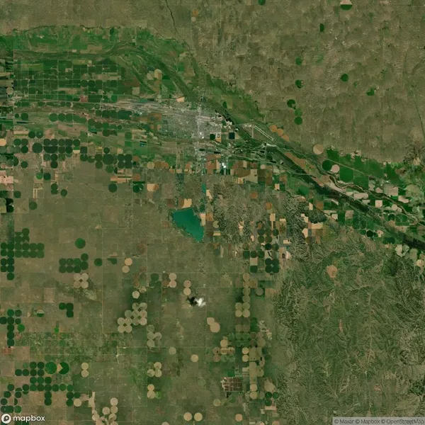 Aerial view of farmland with circular and rectangular fields, a water body, and a town. Various shades of green, brown, and tan indicate different land uses and crops.