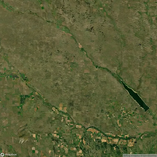 Satellite view of a landscape with patchwork farmland, linear vegetation patterns, and a small lake or reservoir on the right.