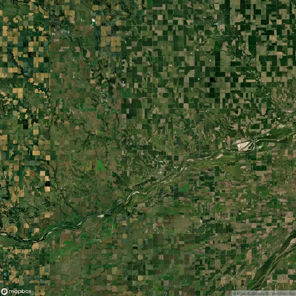 Aerial view of a green landscape with a patchwork of agricultural fields and a river running diagonally through the central area. Sparse forested areas are visible throughout.