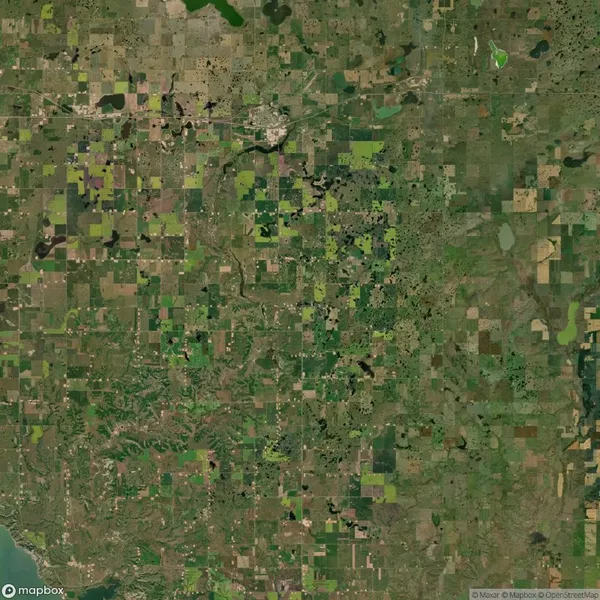 Aerial view of a patchwork landscape with various shades of green and brown fields, intersected by roads and dotted with small bodies of water.