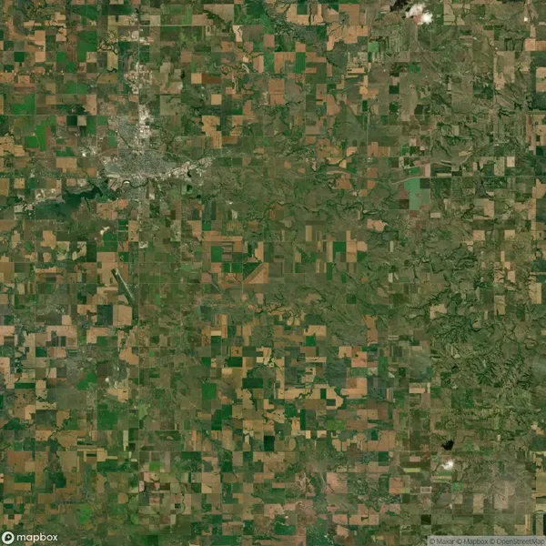 Aerial view of a patchwork landscape with rectangular agricultural fields in varying shades of green and brown, interspersed with small roads and scattered clusters of trees.