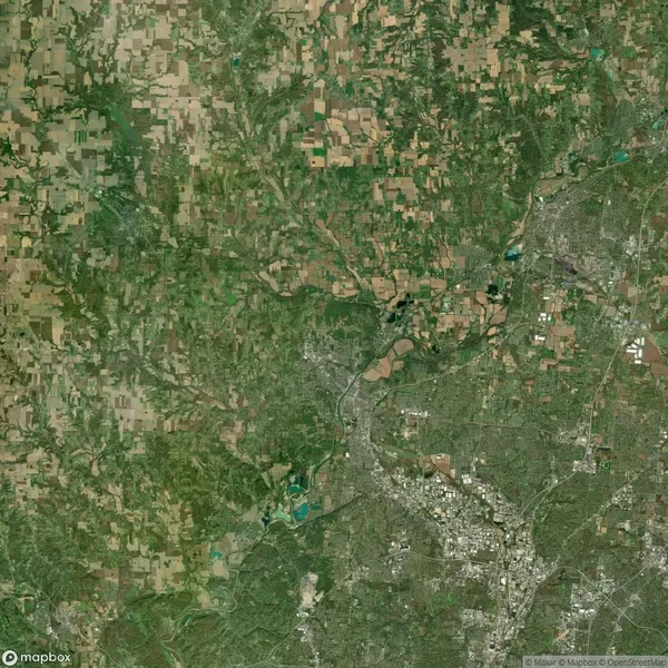 Satellite image showing a mosaic of farmland and urban areas. City center with dense buildings is visible in the lower right, surrounded by green fields and patches of forest.