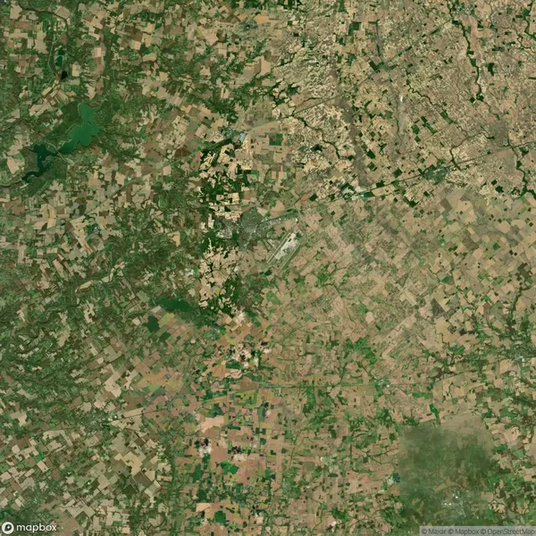 Aerial view of a landscape with a patchwork of agricultural fields in various shades of green and brown, interspersed with patches of forest and a dense urban area in the upper right corner.