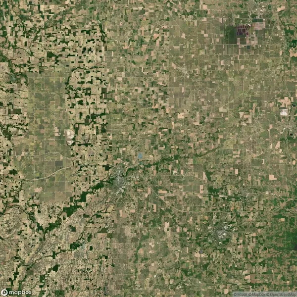 Aerial view of a large patchwork of fields and farmlands with varying shades of green and brown, intersected by roads and small patches of forest.