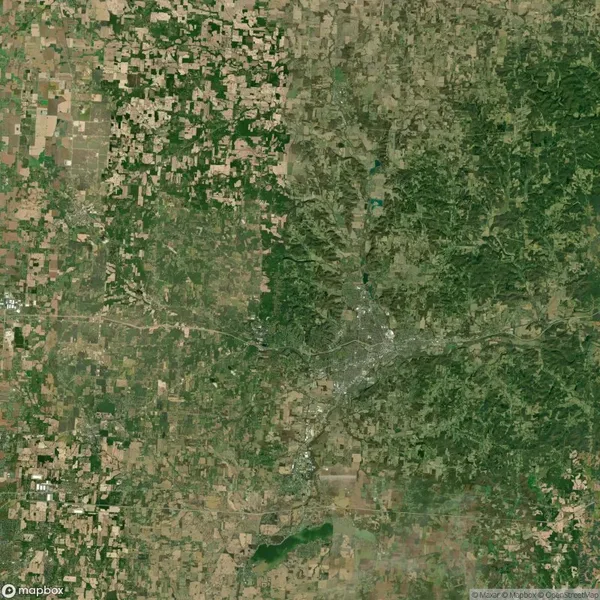 Aerial view of a landscape showing a mix of green forested areas and brown agricultural fields, with roads and a small urban area visible in the center.