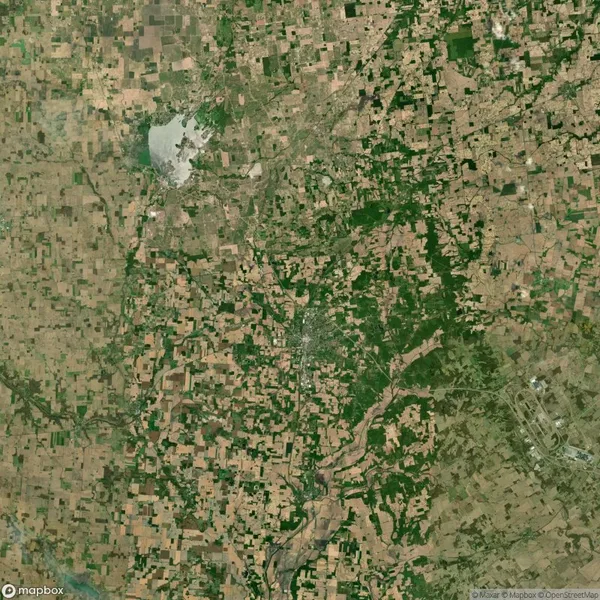 Aerial view of a rural landscape showing a patchwork of farmland, forests, a river, and a large body of water in the upper left corner. Roads and a small urban area are visible.