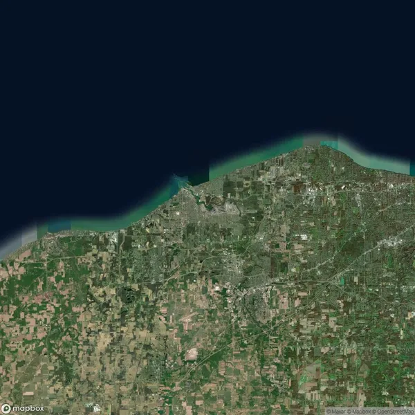 Satellite image of a coastal region with urban areas and agricultural fields. The land meets a body of water at the top, showing a mix of green landscapes and developed regions.