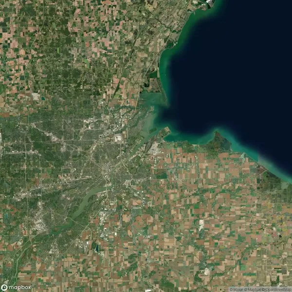 Satellite view of a landscape showing a large body of water meeting land with visible urban and agricultural areas, intersected by various roads and fields.