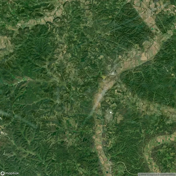 Satellite view of a rural landscape with dense green forests, scattered patches of farmland, and a river running through the terrain. Roads and small settlements are visible.