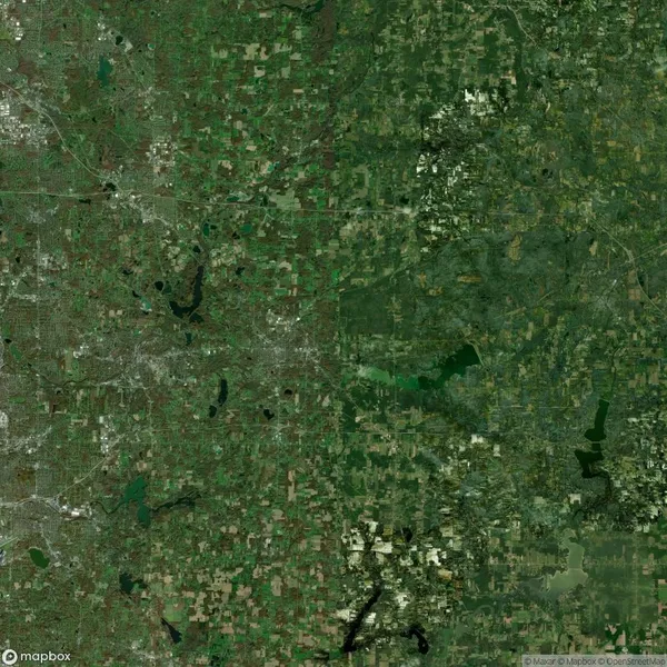 Satellite view of a landscape with a mix of natural green areas and urban development, featuring roads, buildings, fields, and bodies of water.
