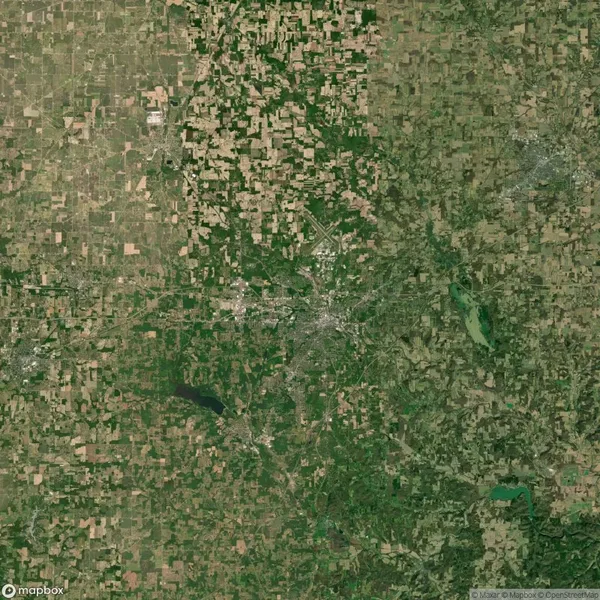 Satellite image of a landscape showing a mix of agricultural fields and urban area at the center, with patches of green and bodies of water scattered throughout.