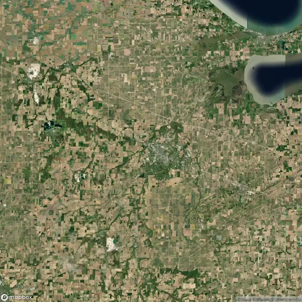 Satellite view of a landscape with a patchwork of agricultural fields and a concentrated urban area near a body of water in the upper right corner.