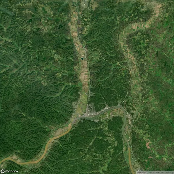 Aerial view of a landscape with winding roads, dense green forests, and scattered patches of farmland. A river flows through the area, surrounded by lush vegetation.