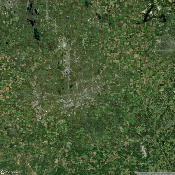 Aerial view of a city surrounded by large expanses of green and brown agricultural fields, with roads and urban areas visible at the center.
