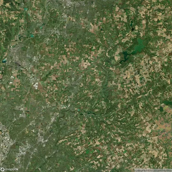 Aerial view of a landscape with a mix of urban areas and agricultural fields, featuring a green river winding through the scene. Roads and patches of forests are also visible.