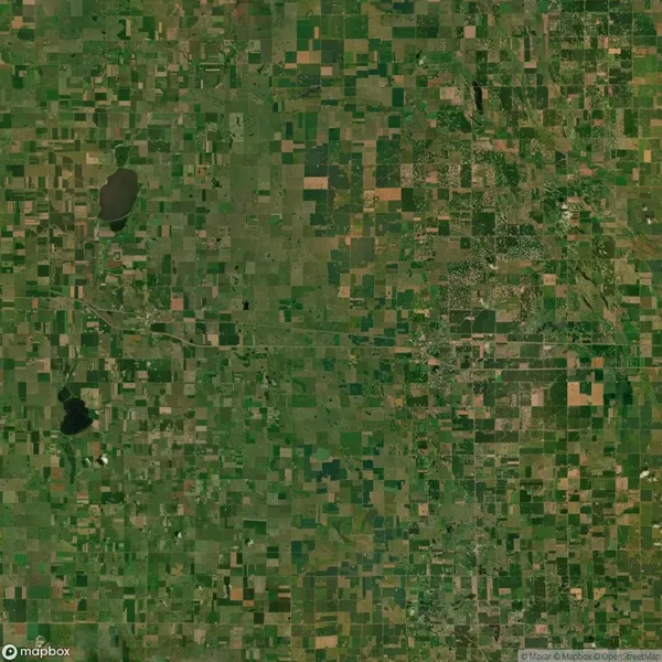 Aerial view of a patchwork of green and brown rectangular fields with scattered bodies of water, resembling a quilt. Roads intersect the landscape, and dense areas hint at settlements.