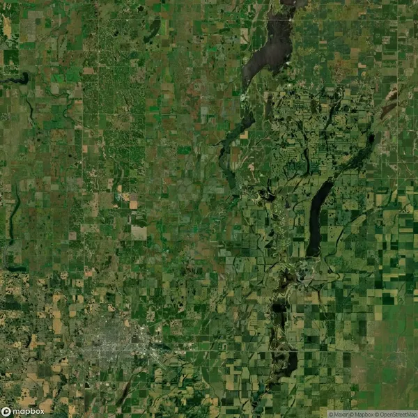 Aerial view of a large, lush green landscape with various shades of green fields, winding rivers, and patches of forested areas. Urban development is visible in the bottom left corner.