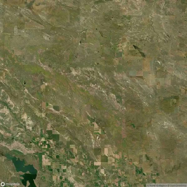 Aerial view of a vast, rural landscape with patchwork fields and sparse vegetation. A body of water is visible in the lower left corner.