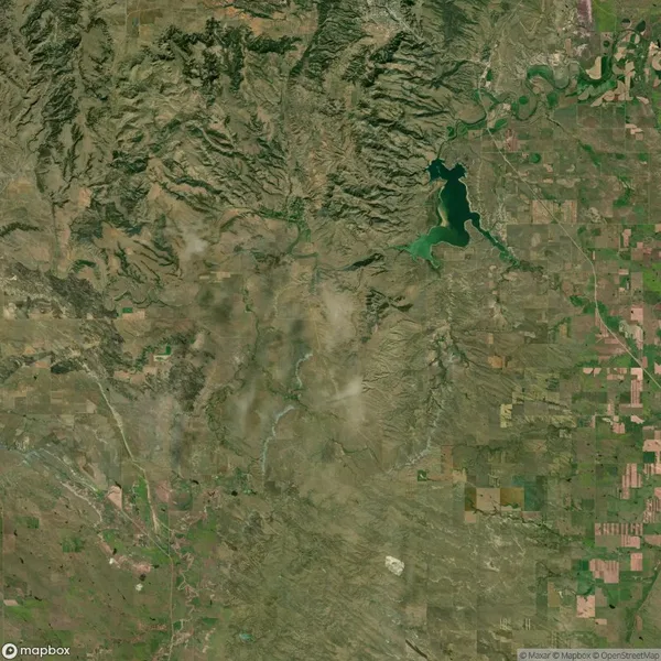 Aerial view of a landscape with farmland patterns on the right, a lake in the upper center, and a rugged, mountainous area on the left. Sparse vegetation is visible.