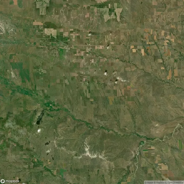 Aerial view of a rural landscape with patchwork fields and a river flowing diagonally. Several clusters of white clouds are visible. The terrain appears flat with scattered vegetation.