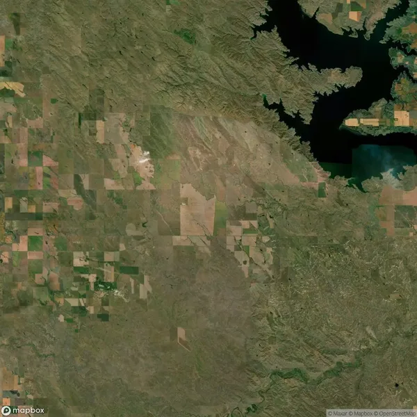 Aerial view of a patchwork landscape with agricultural fields and a body of water in the upper right corner. The area shows a mix of green and brown hues.