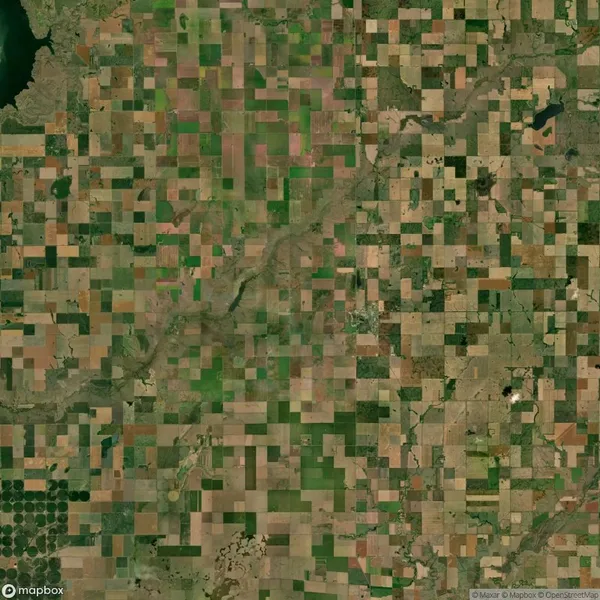 Aerial view of a patchwork landscape featuring agricultural fields in varying shades of green and brown. A river winds through the center, surrounded by a grid-like arrangement of farmland.