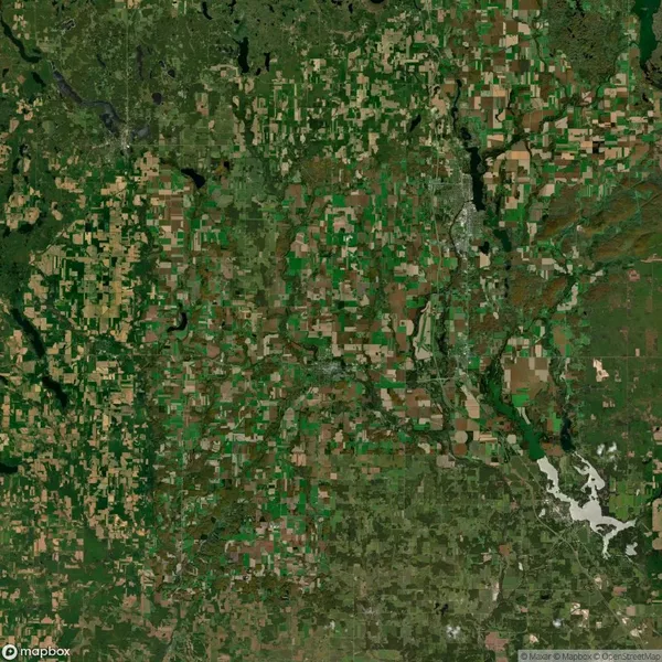 Satellite view depicting a rural landscape with a patchwork of green and brown agricultural fields, interspersed with small wooded areas and a river winding through the scene.