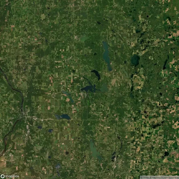 Aerial view of a landscape featuring patches of green fields, small bodies of water, and a network of roads. A river flows in the lower left corner.