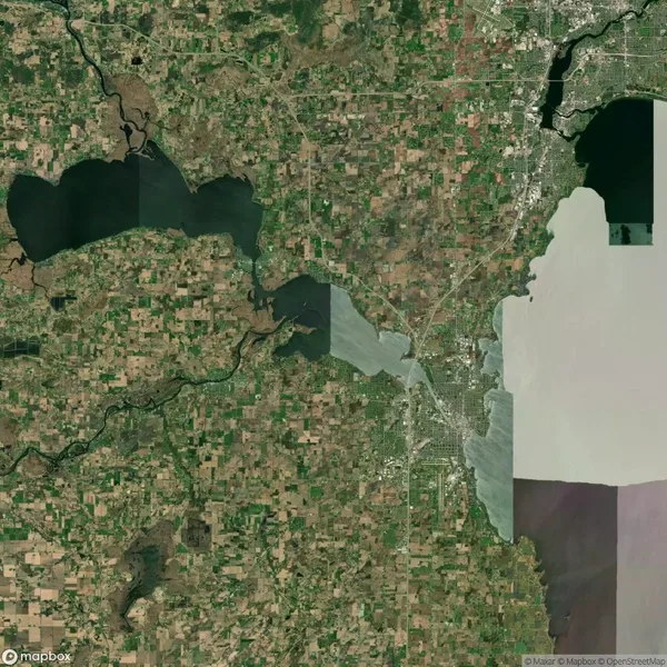 Satellite view of a landscape with lakes, rivers, and a patchwork of agricultural fields. A city area is visible in the upper right quadrant.