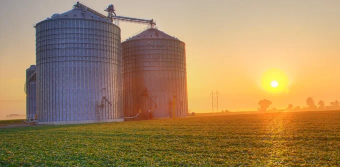 Two large metal silos stand in a field under a clear sky, with the sun setting on the horizon, casting an orange glow over the landscape.