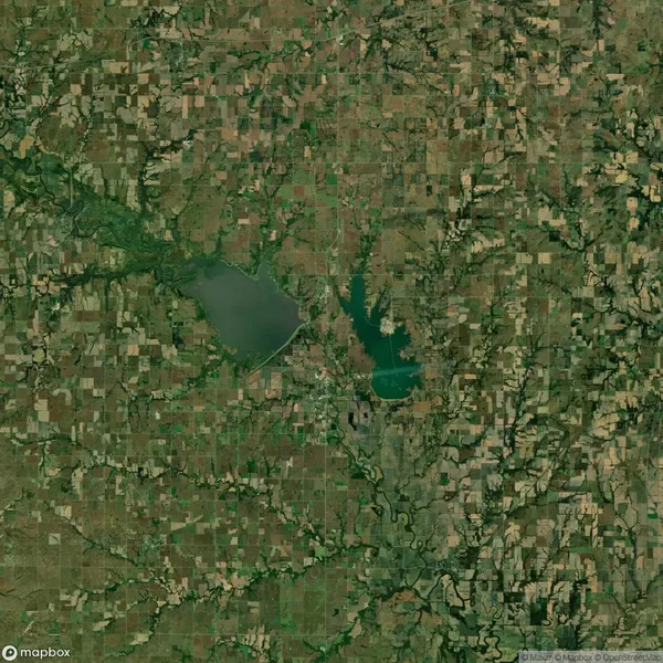 Satellite view of a rural area with a large lake near the center, surrounded by a grid of agricultural fields, patches of forest, and scattered waterways.
