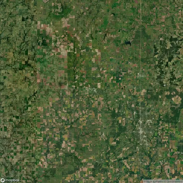 Aerial view of a rural landscape with a patchwork of agricultural fields, scattered wooded areas, and a clustered urban settlement on the lower right.