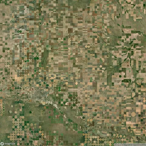 Aerial view of farmland with a patchwork of square and circular fields in various shades of green and brown, intersected by a river and a small town.