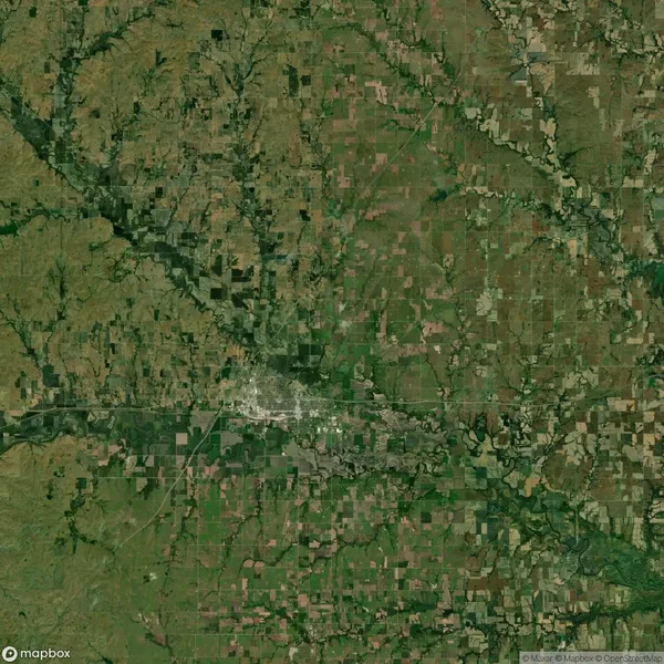 Aerial satellite view of a rural landscape with patchwork farmland and a small city cluster near the center, surrounded by green fields and intersecting roads.