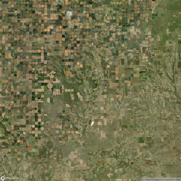 Aerial view of farmland featuring a grid of circular and rectangular crop fields in shades of green and brown, surrounded by patches of natural terrain.