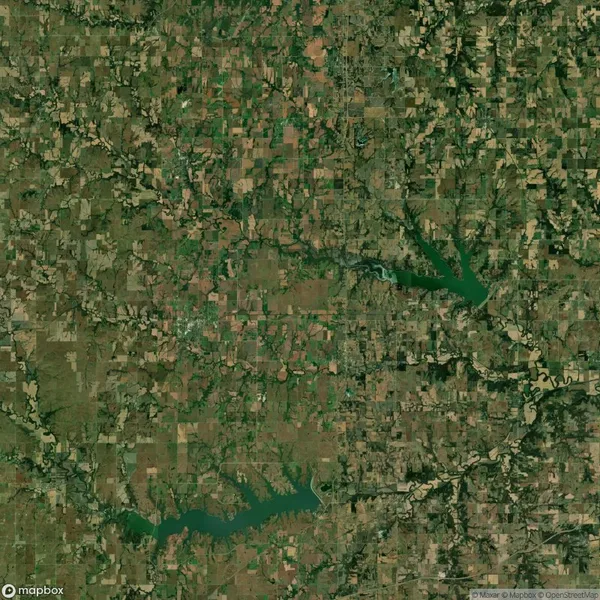 Aerial satellite view of a rural landscape with patchwork fields, clusters of trees, and two large, irregularly shaped green lakes.