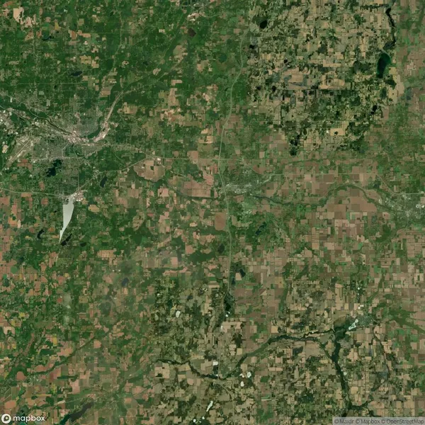 Aerial satellite view of a rural area with a mix of green fields, farmland, and patches of forest surrounding a small urban center in the upper left corner.