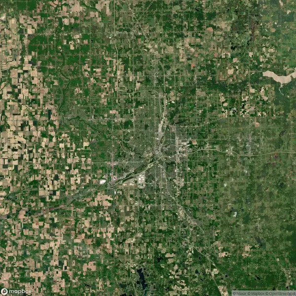 Satellite view of a city surrounded by a patchwork of fields and green areas, showing urban development at the center and rural land on the outskirts.