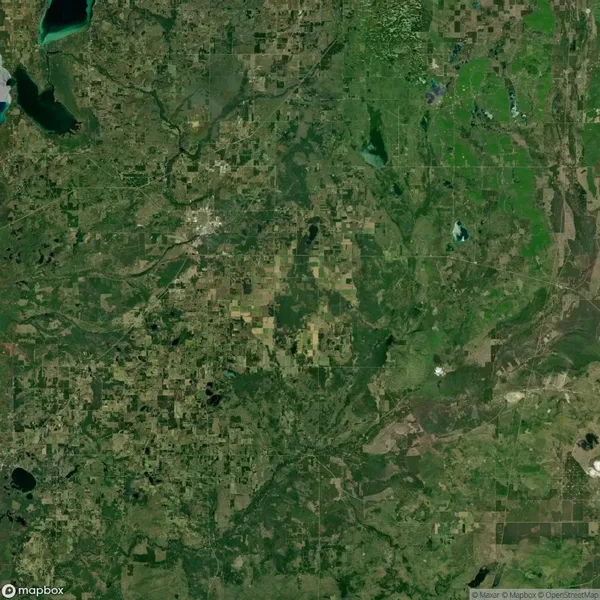 Satellite image showing a mostly green and rural landscape with scattered fields, patches of forest, bodies of water, and a small urban area near the center.