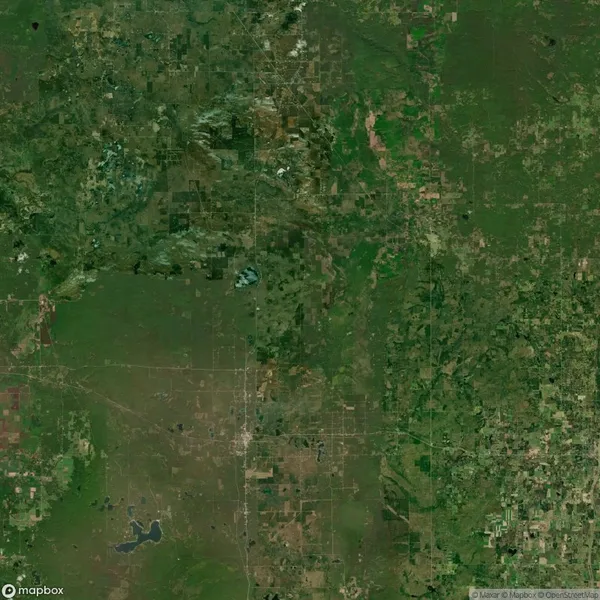 Satellite image showing a patchwork of green and brown land, including fields, forests, roads, and a small lake in the lower left corner.
