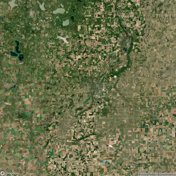 Satellite view of a rural landscape with a patchwork of agricultural fields and a densely built urban area near the center. Scattered bodies of water and green spaces are visible.